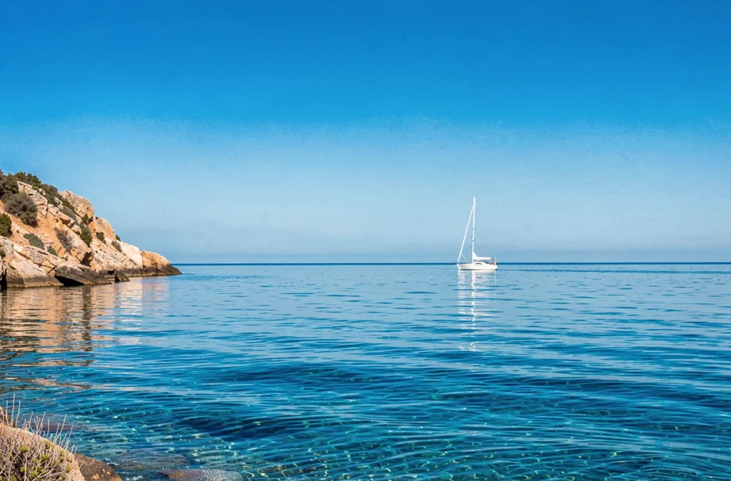 Mer Méditerranée aux teintes azur sous un ciel d'été clair, avec un voilier blanc au loin et des falaises rocheuses